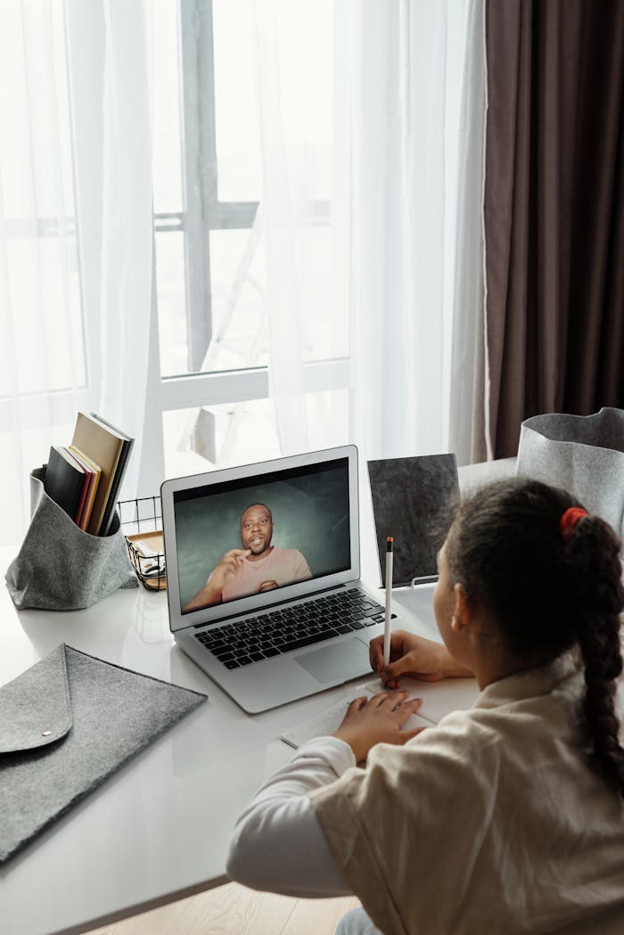 A young girl takes notes during an online class while using a laptop at her study desk.